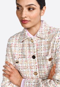 Giacca in tweed con un motivo multicolore e testurizzato su base crema, bottoni neri e dorati, e una camicia colletto rosa sotto.