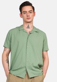 Camisa de manga corta con collar en verde claro, tejido texturizado con patrones sutiles, cierre de botones y corte relajado.