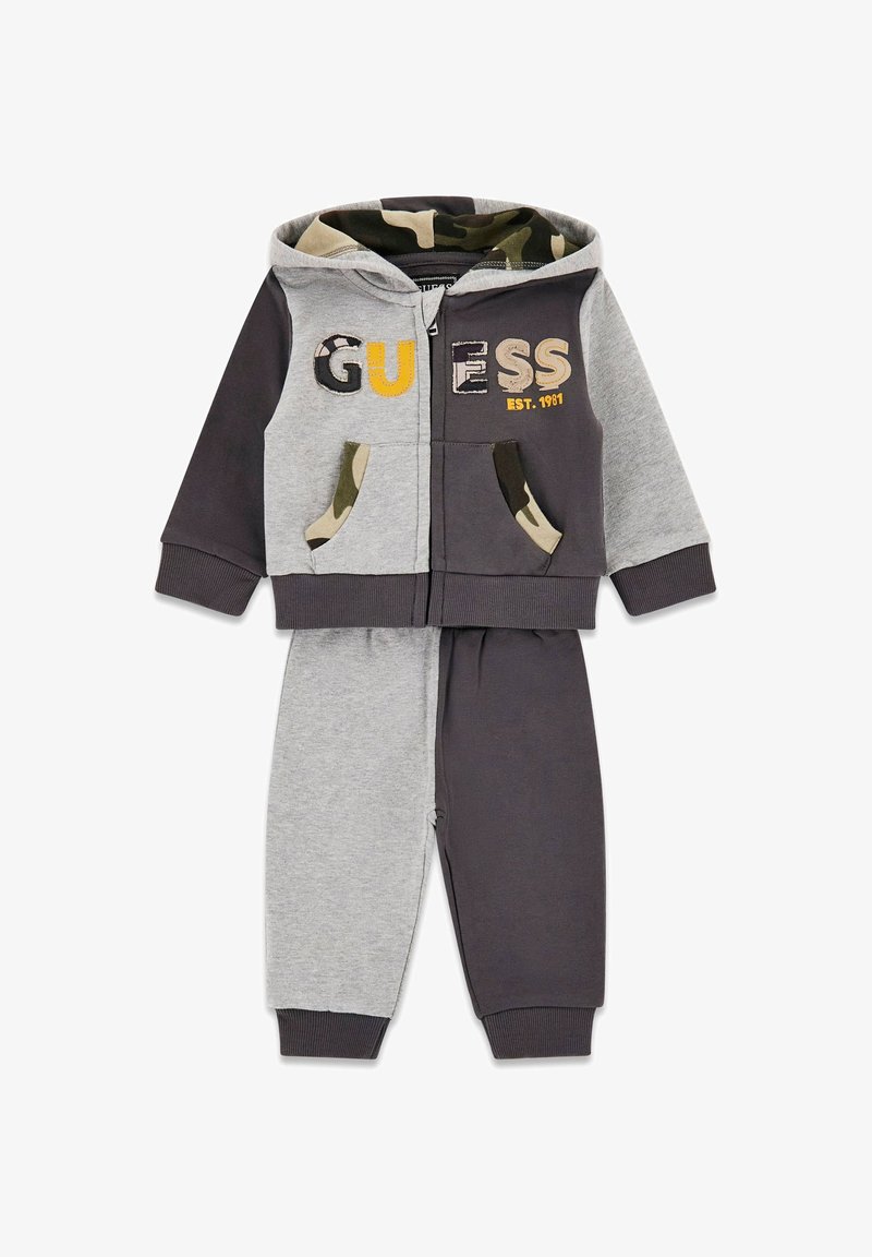 Grau und dunkelgrauer zweiteiliger Trainingsanzug mit einer kapuzenbesatz aus Tarnmuster, der das Wort "GUESS" in verschiedenen Farben auf der Vorderseite und an den Taschenakzenten zeigt.