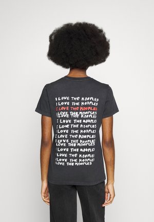 Osoba z ciemnymi kręconymi włosami w czarnej koszulce z powtarzającym się białym i jednym czerwonym napisem „I LOVE THE KOOPLES” na plecach.