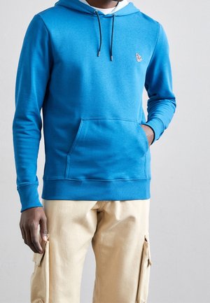 Sininen huppari, jossa on huppu, puuvillamateriaalia, etutasku ja kiristysnyöri. Rinnassa pieni logo. Yhdistetty beigenvärisiin cargo-housuihin.