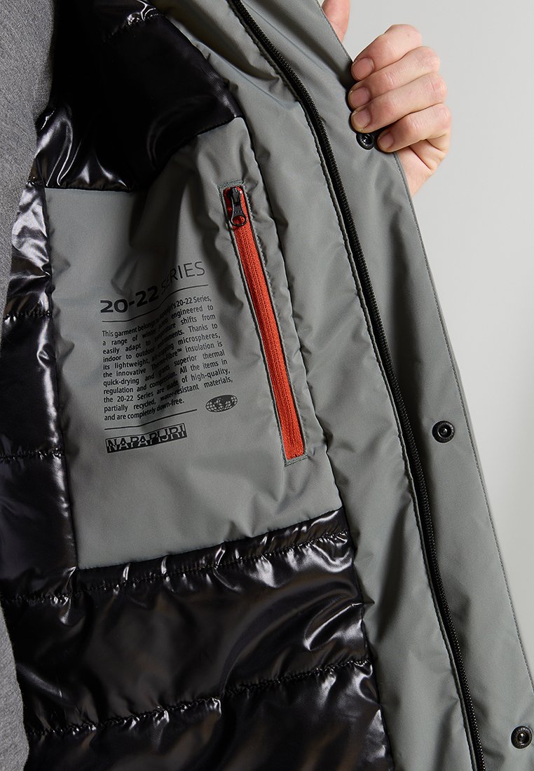 Interno di un piumino grigio con una sezione rivestita di nero, una tasca con cerniera e dettagli arancioni, e etichette stampate che descrivono il prodotto.