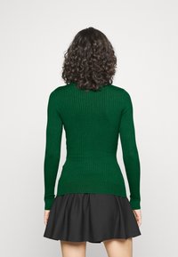 Femme aux cheveux bruns bouclés portant un pull vert côtelé ajusté et une jupe noire évasée, vue de dos sur un fond blanc.