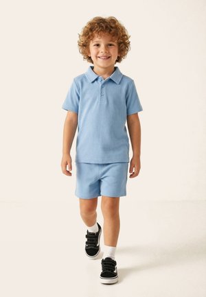 Ragazzo giovane con capelli ricci che indossa una polo azzurro chiaro e pantaloncini, cammina avanti con un sorriso su uno sfondo neutro.