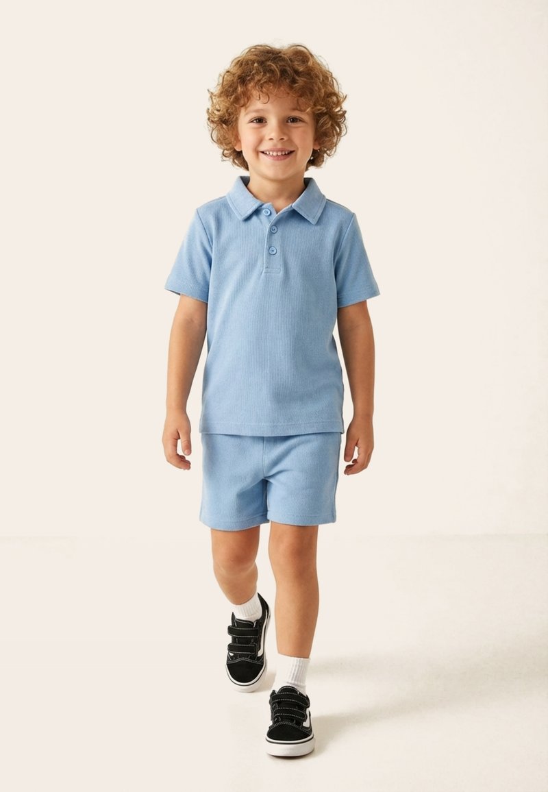 Jeune garçon aux cheveux bouclés portant un polo bleu clair et un short, marchant en avant avec un sourire sur un fond uni.