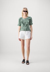 Top corto con maniche a sbuffo di colore verde con fantasia, abbinato a pantaloni corti bianchi con orlo sfrangiato e mule nere aperte davanti.