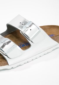 Sølvmetallic slide-sandal med to justerbare spænder, korkbund og tekstureret gummisål. Har Birkenstock-branding i blå.