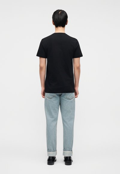 Μαύρο βαμβακερό t-shirt με κοντά μανίκια, σε προβολή από πίσω. Συνδυασμένο με τζιν παντελόνι ανοιχτό μπλε και μαύρα παπούτσια. Απλός, καθημερινός σχεδιασμός.