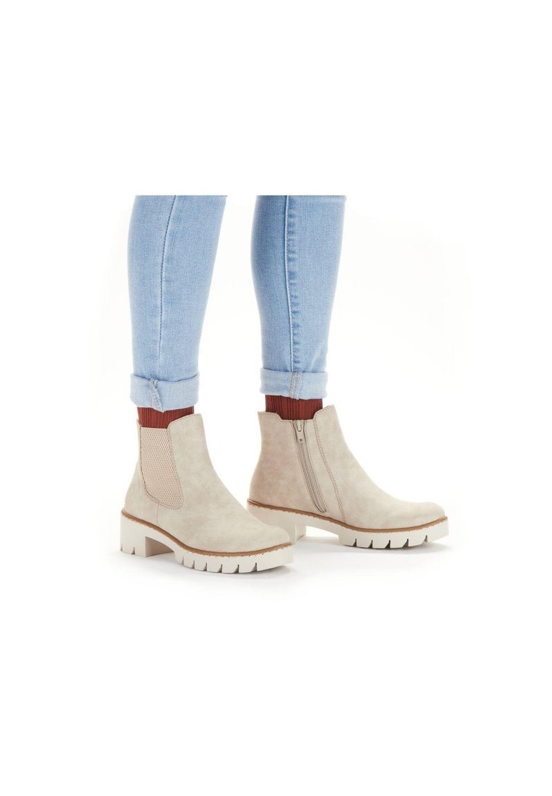 Bottines beiges en suède avec des panneaux latéraux en élasthanne, une fermeture éclair sur le côté et une épaisse semelle blanche, associées à un jean bleu clair retroussé.