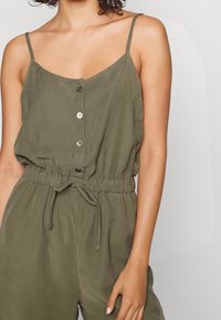 Combinaison vert olive avec fines bretelles spaghetti, fermeture avant à boutons, taille élastique et cordon de serrage. Tissu lisse et léger.