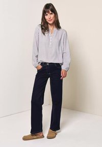 Mujer de pie y sonriendo, con una blusa blanca de rayas, vaqueros oscuros de pierna ancha y zapatos color beige sin cordones sobre un fondo liso.