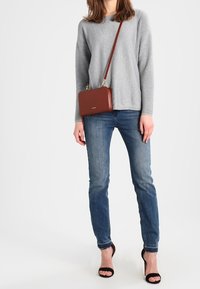Graues geripptes Langarmoberteil, blaue Denim-Jeans, braune Leder-Crossbody-Tasche, schwarze Sandalen mit offenem Zeh, vor einem neutralen Hintergrund stehend.