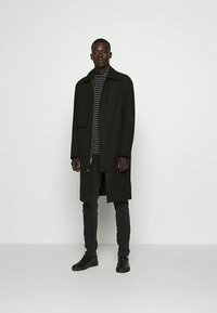 Visok muškarac koji nosi crni dugi kaput preko crno-sivog prugastog rolke, crne hlače i crne cipele, stoji ispred jednostavne svijetle pozadine.