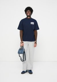 Marine T-skjorte med logo, grå joggebukser, blå håndveske med logo, og blå sko med tre striper, stående mot en ensfarget bakgrunn.