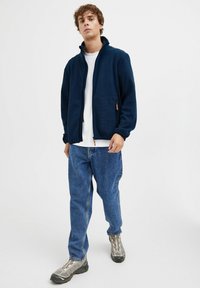 Marineblå zip-up jakke med en teksturert overflate, to frontlommer og kontrasterende beige detaljer. Brukt over en hvit t-skjorte og denim jeans.