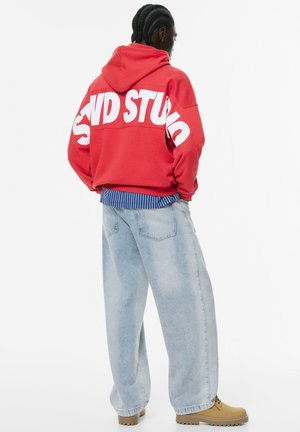 Man with braided hair wearing red hoodie with bold white "SND STUDIO" text, loose light blue jeans, striped shirt, and tan boots standing sideways.