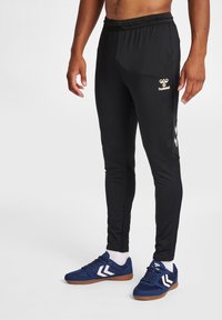 Schwarze Sporthose mit schlanker Passform, elastischem Bund und goldenem Logo auf der linken Hüfte. Kombiniert mit blauen Sportschuhen mit weißen Akzenten.