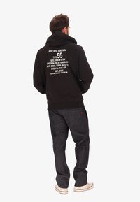 Felpa nera con testo bianco sulla schiena, abbinata a pantaloni in denim scuro e scarpe di colore chiaro. La felpa ha un ampio cappuccio e polsini a coste.