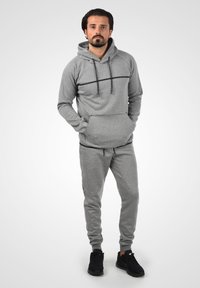 Grå hættetrøje med en frontlomme, kontrasterende sort vandret stribe, parret med matchende grå træningsbukser og sorte sneakers.