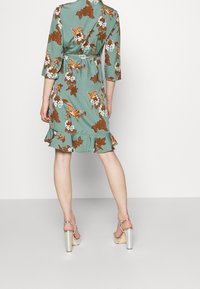 Vihreä kukallinen mekko, jossa on röyhelöhelma, kolmanneksen pituiset hihat ja vyötäröllä vyö, jossa on ruskeita ja valkoisia kukkakuoseja.