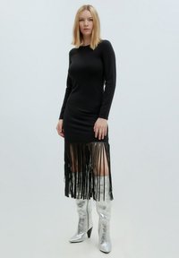 Schwarzes Langarmkleid mit Fransendetails am Saum, aus geripptem Stoff; kombiniert mit silberner, metallischer Knielanger Stiefeln.