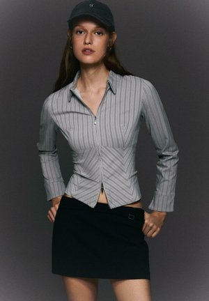 Donna che indossa una maglia grigia a righe con zip, mini gonna nera e cappello nero, in piedi con le mani sui fianchi davanti a uno sfondo grigio scuro.