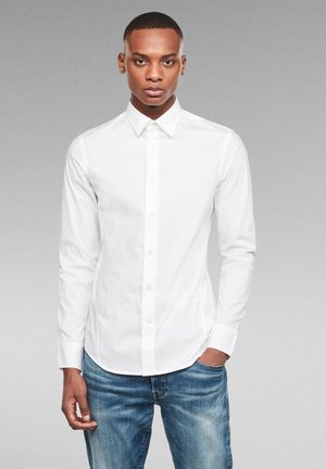 Camisa blanca de botones con cuello puntiagudo, mangas largas y un diseño ajustado, combinada con jeans azules. Textura suave, sin patrones.