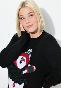 Frau mit kurzem blonden Haar, die einen schwarzen Weihnachtspullover mit einem Schneemann und "Frohe Weihnachten"-Text trägt, während sie ihren Kopf auf ihrer Hand abstützt.