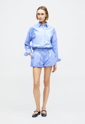 Naine seisab, kandes helesinise triibulise pikkade varrukatega nööpidega särgi ja sobivaid lühikesi pükse, jalas mustad Mary Jane stiilis kingad, taust valge.