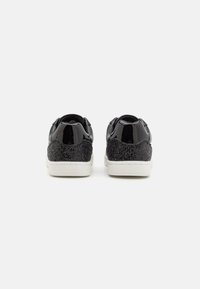 Zapatillas de charol negro con un talón texturizado de purpurina negra, suela de goma blanca y diseño de parte trasera redondeada.