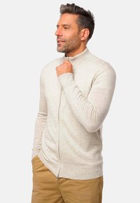 Un sweat à zip gris clair avec un col haut, des poignets côtelés et une texture lisse, porté sur un pantalon beige.