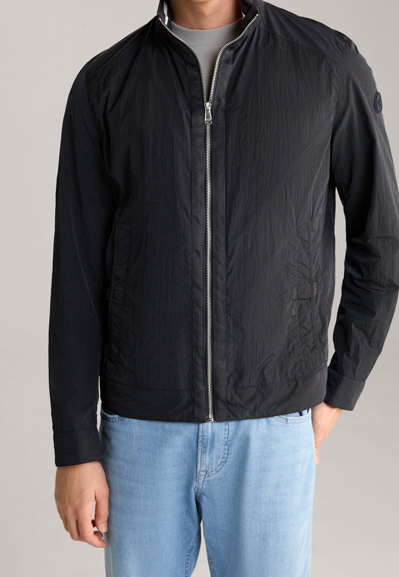 Man draagt een zwarte jas met rits over een grijs shirt en lichtblauwe spijkerbroek, staand tegen een effen achtergrond.