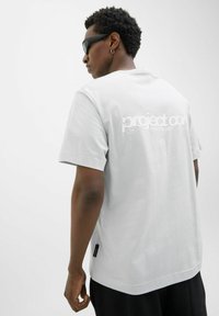Lys grå bomulds T-shirt med korte ærmer, med et stort hvidt logo på ryggen, standard pasform, rund halsudskæring og en sidelabel detalje.