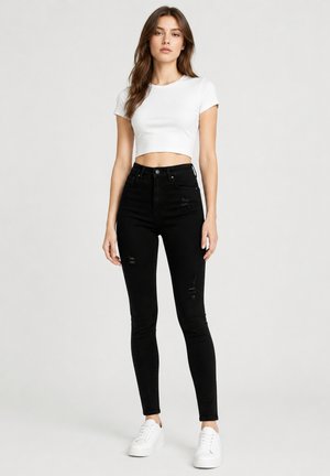 Ung kvinde står iført en hvid crop top, højtaljede sort revnede skinny jeans og hvide sneakers mod en ensfarvet lys baggrund.