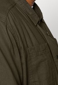 Camicia verde oliva con bottoni, colletto a punta, taschino frontale e dettagli in tessuto testurizzato. Cuciture visibili e bottoni neri.
