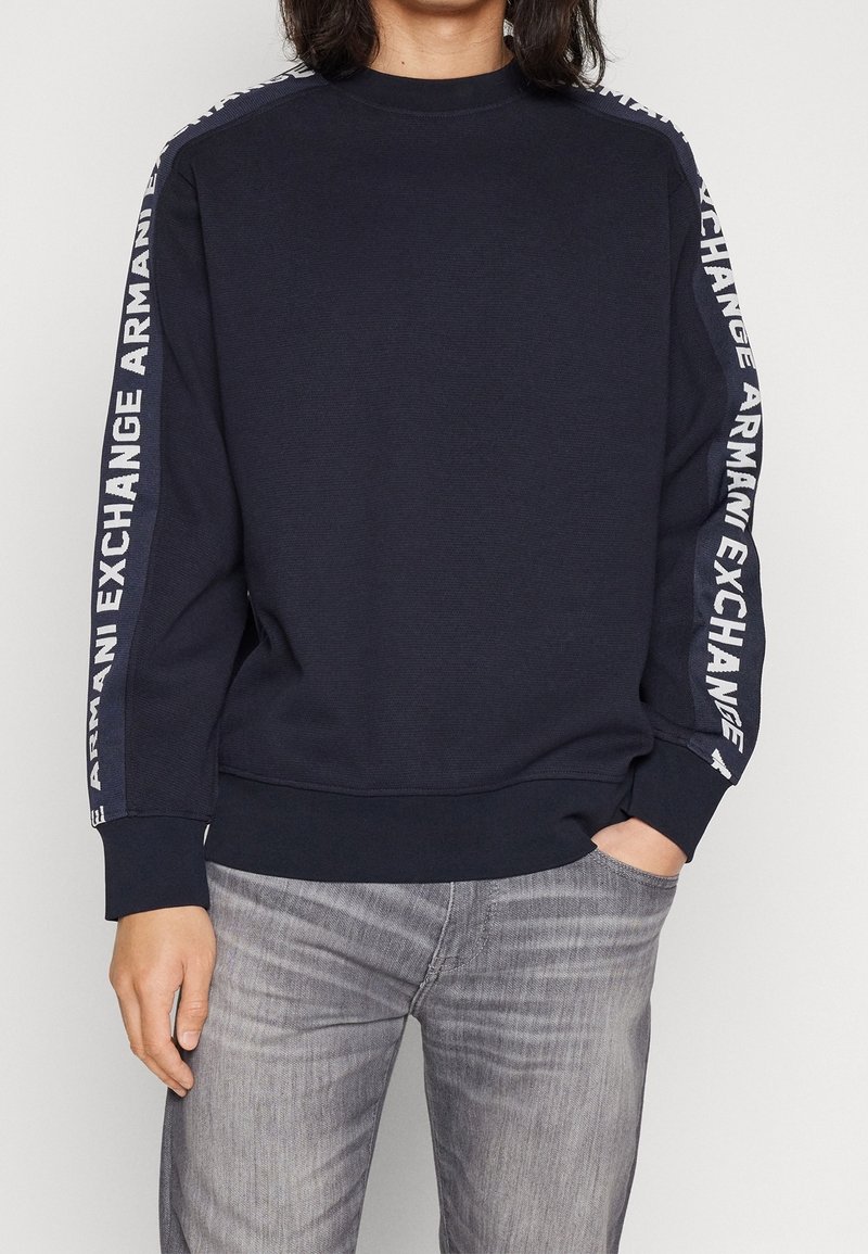 Personne portant un sweat-shirt bleu marine avec le texte « Armani Exchange » sur les deux manches et un jean gris délavé sur un fond uni.