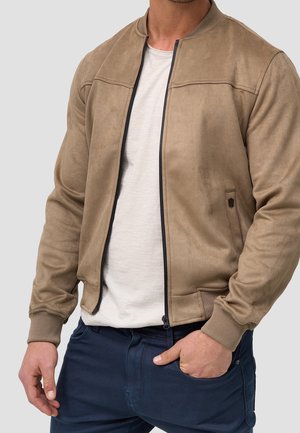 Mann trägt eine beige Bomberjacke mit Reißverschluss über einem weißen T-Shirt und marineblaue Hosen, mit einer Hand in der Tasche und der anderen locker an der Seite.