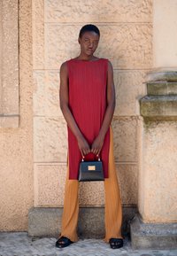 Vestido vermelho plissado sem mangas combinado com calças plissadas bege e sandálias chunkys pretas. Bolsa preta com fecho dourado segurada nas mãos.