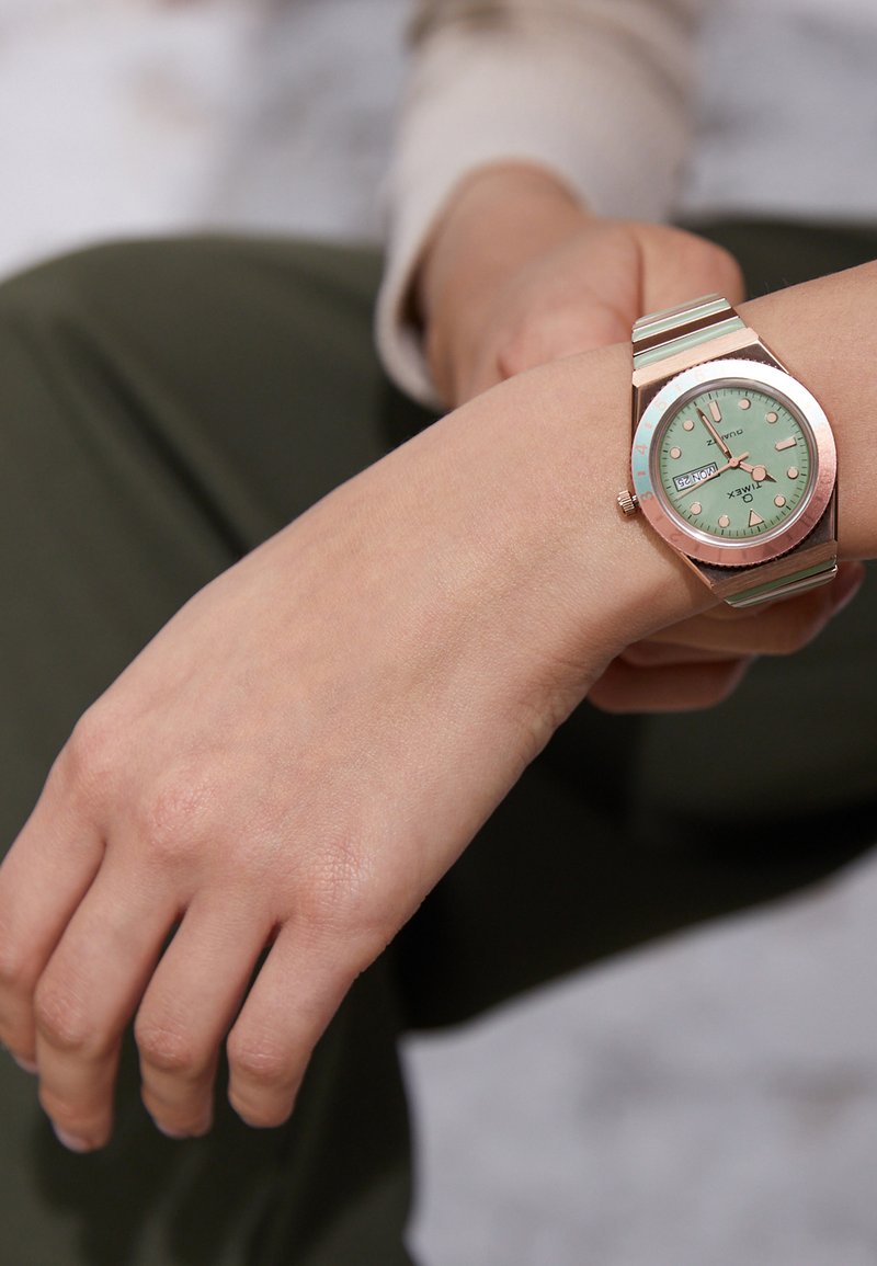 Montre dorée avec un cadran vert menthe, bracelet à rayures argentées et boîtier rond. Elle présente des marqueurs d'heure numériques et un affichage de la date.