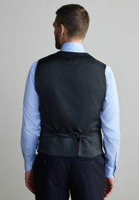 Schwarze Weste mit glatter Textur, figurbetontem Design und verstellbarem Riemen im Rücken, getragen über einem hellblauen Hemd.