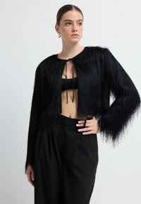 Kobieta w czarnej, teksturowanej, skróconej kardiganie na czarnym crop topie i czarnych, wysokich spodniach, pozująca z ręką na biodrze na białym tle.