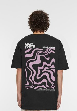 Persona con cabello rizado lleva una camiseta negra con un patrón abstracto ondulado en rosa y negro y el texto "Lost Youth" y "Find beauty in the chaos" en la espalda.