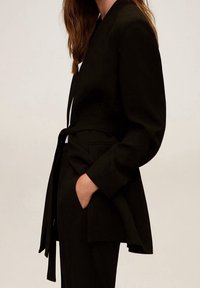 Sorte skræddersyet blazer med lange ærmer, bælte i taljen og en glat tekstur. Har en sjal krave og sidelommer.