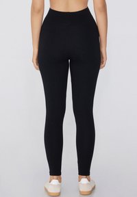 Leggings noirs avec une taille haute et une texture lisse et extensible, ajustées étroitement sur les jambes, portées avec des chaussures claires.