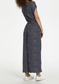 Jumpsuit floral bleu foncé avec des manches courtes, taille élastique et jambes larges, associé à des baskets blanches.