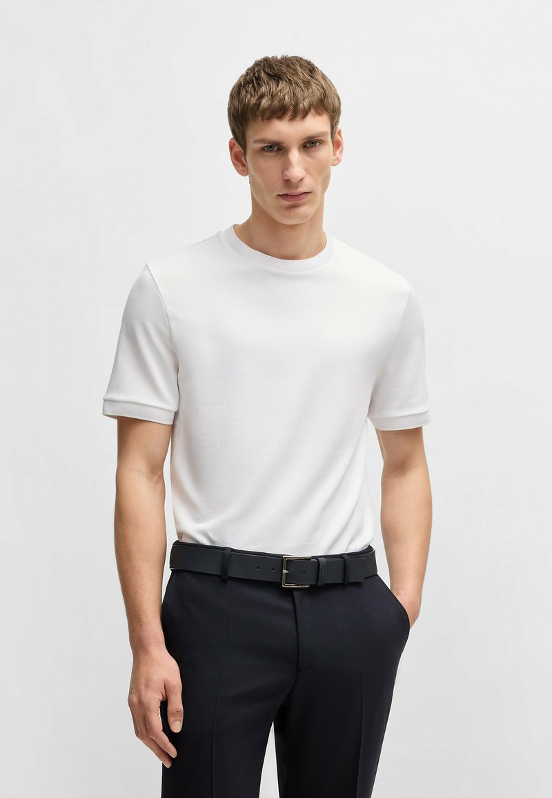 T-shirt bianco a maniche corte con collo rotondo, realizzata in tessuto testurizzato, abbinata a pantaloni scuri su misura e una cintura nera con fibbia rettangolare.