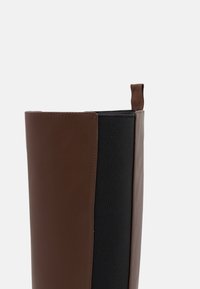 Bota alta de cuero marrón con panel elástico negro en el lateral y lengüeta marrón, sobre un fondo liso gris claro.