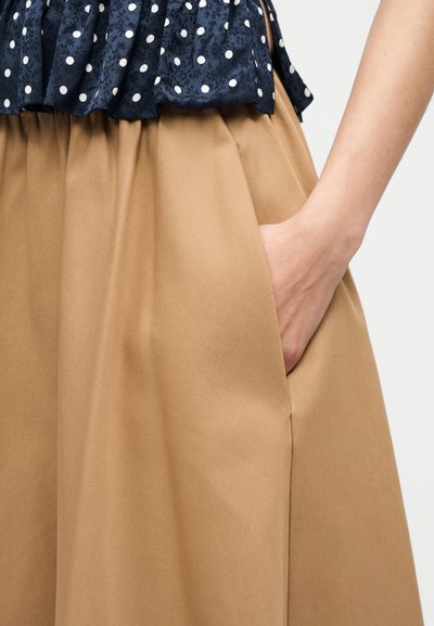 Χέρι γυναίκας που ξεκουράζεται στην τσέπη μιας φούστας ψηλόμεσης σε καμηλό χρώμα, σε συνδυασμό με μπλε πουά τοπ με ναυτικό μπλε χρώμα.