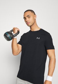 Černé sportovní tričko s texturovaným mřížkovaným vzorem, sivým logem na hrudi, nošené osobou, která drží kettlebell.
