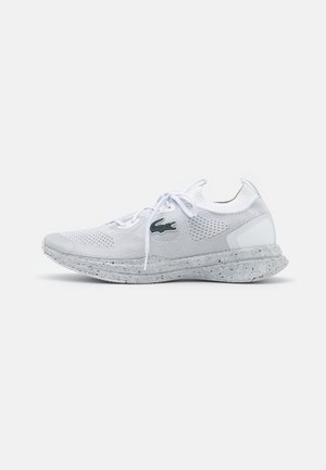 Chaussure de sport blanche en maille, avec semelle mouchetée, lacets à l’avant et logo crocodile sur le côté, montrée de profil sur un fond uni.